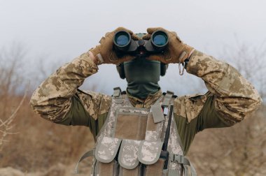 Kamuflajlı bir asker dürbünü tutuyor. Askeri adamın elinde dürbün var. Sahadaki asker