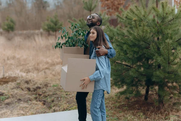 A happy young family is moving into a house. A black man and woman are holding cardboard boxes for moving, smiling. moving boxes