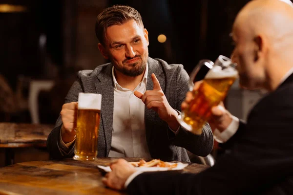 adult friends sitting with beer mugs in a pub. Two cheerful guys are drinking draft beer, celebrating meeting and smiling.
