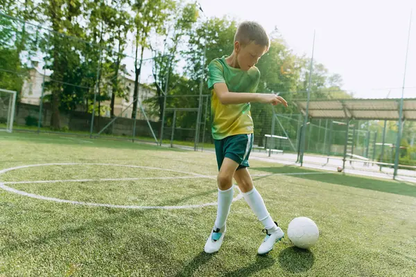 Yeşil üniformalı bir futbolcu çocuk futbol sahasında topla koşuyor.