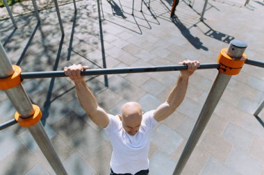 Beyaz tişörtlü kel bir adam açık havada yatay bir barda çalışıyor.