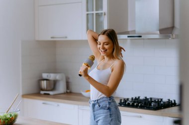 Evde şarkı söyle. Genç bir ev hanımı, rahat ve modern bir mutfakta dans edip şarkı söyler. Genç bir kadın mutfağında mikrofona şarkı söyleyerek eğleniyor.