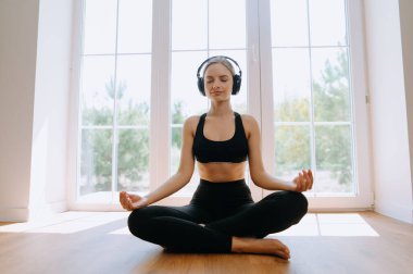 Güzel ve sportif kız sabahları evde meditasyon yapıyor ve kulaklıkla müzik dinliyor. Pencerenin kenarındaki sabahında. Meditasyon için lotus konumu