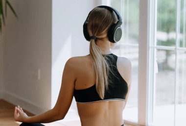 Güzel ve sportif kız sabahları evde meditasyon yapıyor ve kulaklıkla müzik dinliyor. Arkadan bak. Pencerenin kenarındaki sabahında. Meditasyon için lotus konumu