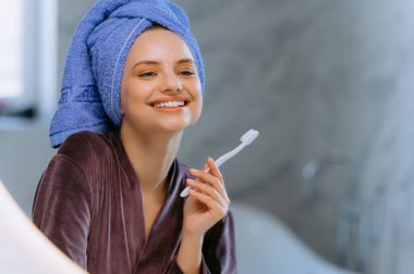 Diş fırçası tutan, dişlerini fırçalayan genç bir kadın aynaya bakıyor. Sabah banyoda ağız temizleme, diş bakımı rutini. Beyaz dişler
