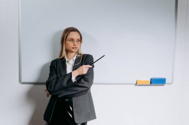 Sınıfta karatahtanın önünde gülümseyen öğretmen. Genç bayan öğretmen elinde kalemle beyaz bir yazı tahtasının arkasında konuşuyor.