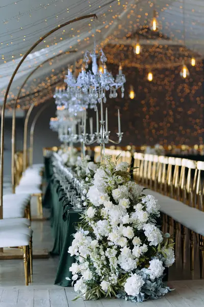Lüks düğün ziyafeti salonu. Düğün kutlaması için hazırlanmış bir masa. Beyaz buketler, şeftali çiçekleri, klasik dekorlar. Beyaz parlak oda, bir gül kemeri