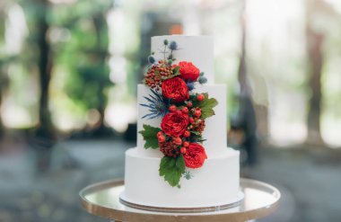 Pasta beyaz krema ile kaplıdır ve masada servis edilen kırmızı bir çiçekle süslenir. katlı düğün pastası