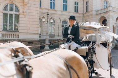 Resmi giyinmiş bir kadın, kendine güvenen bir şekilde at arabasında oturur. Arkaplanda büyüleyici bir şehir deneyimini vurgulayan süslü bir bina yer alıyor..