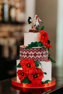 Zarif düğün pastası gösterileri çiçekli elementler ve eşsiz süslemeler. Birleşmeyi sembolize etmek için koyu kırmızı çiçekler, desenli bantlar ve leylekler..