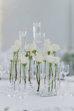 A beautifully arranged table featuring white roses in glass vases, fine china, and cutlery. The setting is perfect for a wedding or formal event with a chic ambiance. clipart