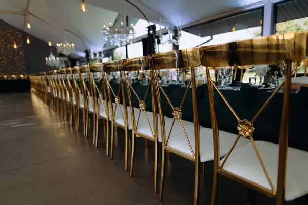 Misafirler için uzun sıra altın koltukların yer aldığı şık bir ziyafet sahnesi. Karanlık örtülerle süslenmiş masalar lüks bir yemek atmosferi yaratır..