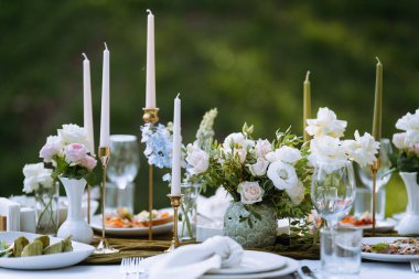 Açık hava yemek masasında zarif çiçekli süsler ve parlayan mumlar var. Lezzetli yemek tabakları cam eşyalar arasında güzelce yerleştirilmiştir..