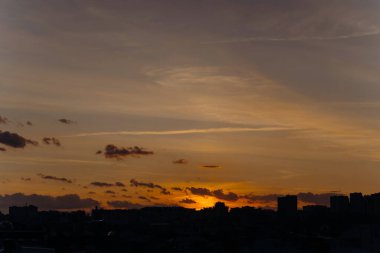 Şehir günbatımında ufukta canlı renkler ve siluetler sergileniyor. Güneş şehir binalarının arkasında batarken bulutlar büyüleyici bir atmosfer yaratıyor..