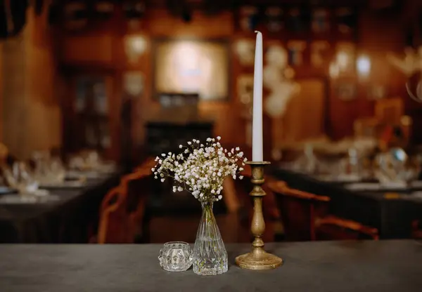 Dekoratif masada bir vazo narin çiçeğin yanında ince bir mum bulunur. Arka planda sıcak, kırsal bir yemek atmosferi ve karanlık masalar var..