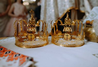 Süslü iki altın taç zarif bir şekilde bir masanın üzerinde oturuyor, karmaşık tekstil ile çevrili, bir düğün töreninde aşkı ve birlikteliği simgeliyor.