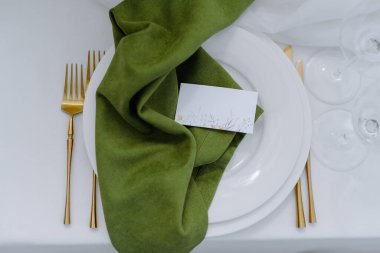 Altın çatallı, parlak yeşil peçeteli ve çiçekli detaylarla süslenmiş güzel bir masa düşünün..