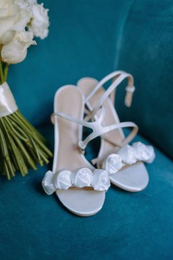 Süslü beyaz sandaletler kumaş güllerle süslenmiş, güzel bir gelin buketinin yanında sergileniyor..