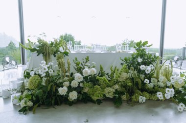 Beyaz ve yeşil çiçeklerle süslenmiş güzel bir masa, bardaklar ve doğa manzarası, sakin bir ortam için mükemmel..