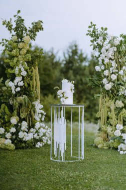 Çağdaş bir standın üzerinde beyaz bir pasta sergilenen çarpıcı bir düğün, taze çiçekler ve yeşilliklerle süslenmiş, sakin bir arka planda..