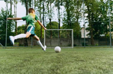 Bir çocuk, yemyeşil bir sahada topa güçlü bir tekme atarak futbol yeteneklerini sergiliyor. Arkasında açık havada bir kale direği bulunuyor..