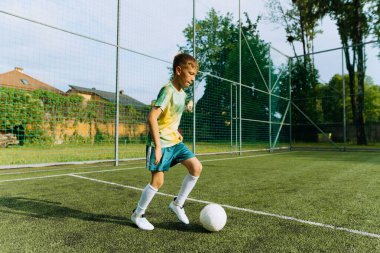 Genç bir çocuk yemyeşil bir arazide futbol topu sürüyor, gündüz saatlerinde açık hava sporlarında yeteneklerini sergiliyor..