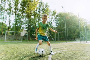 Genç bir çocuk sahada futbol becerilerini gösteriyor, çevikliğini gösteriyor ve sıcak güneş ışığına odaklanıyor..