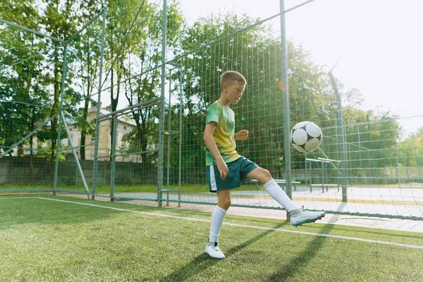 Genç bir çocuk, yeşil bir kale direği ve güneş ışığıyla ıslanmış bir ortamda, ayağındaki topu dengeleyerek futbol becerilerini geliştiriyor..