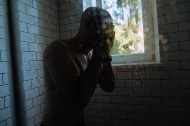 Bir adam, parlak bir duşta dururken yüzünü yıkarken görülüyor, pencereden süzülen doğal ışık, sakin bir atmosfer yaratıyor..