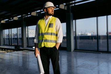 Bir inşaat yöneticisi, tamamlanmamış bir ofiste elinde planlar, güvenlik yeleği ve kasketi ile projeyi yönetmeye hazır bir şekilde özenle duruyor..