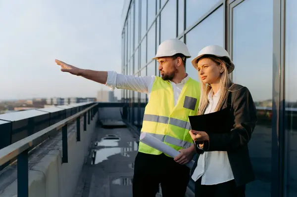 Bir inşaat yöneticisi ve bir meslektaşı, balkondan bir inşaat alanını incelerken....