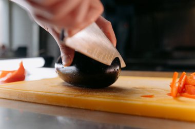 Bir aşçı patlıcanları ustalıkla dilimler. Kesme tahtasının üzerinde bir bıçakla. Yanında kırmızı dolma biber parçaları..