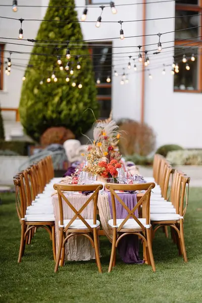 Güzel bir şekilde düzenlenmiş açık hava yemek masası misafirleri bekliyor, canlı çiçekler ve ortam yaylı ışıklarla süslenmiş, kutlamalar için rahat bir ortam yaratıyor..