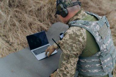 Kamuflaj teçhizatı kullanan bir askeri ajan, saha ortamında veri analizi yaparken bir dizüstü bilgisayara odaklanır..