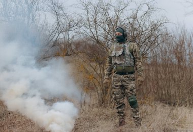 Kamuflaj elbisesi giymiş bir asker dumanın ve yoğun yeşilliklerin arasında duruyor. Uzak bir orman ortamında harekete geçmeye hazırlanıyor..