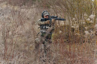 Taktik teçhizatla donatılmış bir asker eğitim ya da çatışma sırasında hazırlık ve hassasiyet gösteren bir tüfeği doğal bir ortamda çömelir ve nişan alır..