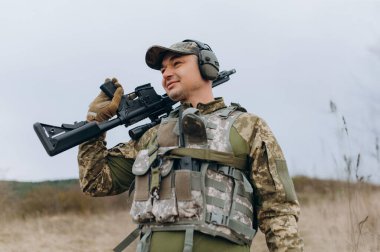 Kamuflaj kıyafeti giymiş bir asker, elinde tüfekle doğal bir ortamda güvenle ayakta duruyor. Sahne, barışçıl bir arkaplan arasında bir hazırlık ve kararlılık hissi yakalıyor..