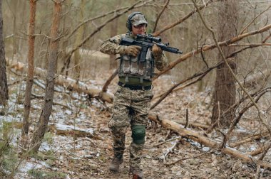 Silahlı ve taktik teçhizatlı bir asker ormanlık bir alanda gezinerek açık havada hazırlık ve eğitim gösteriyor..