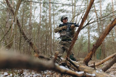 Yoğun bir ormanda silah tutarken kamuflajlı bir asker hazır olduğunu ve taktiksel bir ortama odaklandığını gösteriyor..