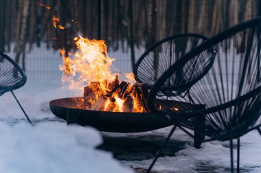 A warm and inviting fire pit surrounded by snow and modern chairs, perfect for winter gatherings and relaxation.
