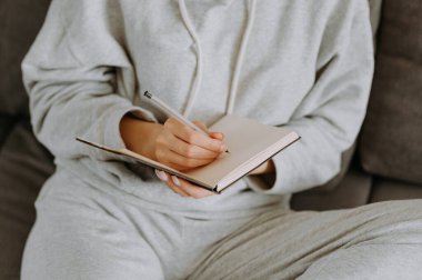 Gri elbiseli bir kadın kanepede rahat bir şekilde otururken bir deftere yazar..