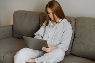 Gri kapüşonlu genç bir kadın rahat bir şekilde kanepede oturuyor, laptopuna odaklanmış, rahat bir atmosferi kucaklıyor..