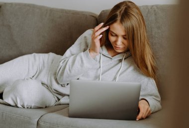 Genç bir kadın rahat bir yerde laptopuna odaklanmış bir şekilde kanepede uzanıyor..