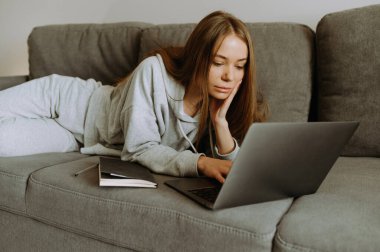 Gündelik giysiler içinde genç bir kadın kanepeye uzanıyor, yoğun bir şekilde dizüstü bilgisayarına odaklanmış bir şekilde, evinde rahat bir çalışma alanı sergiliyor..