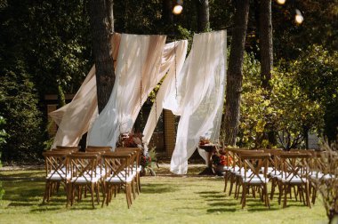 Güzel bir açık hava düğün töreni, akıcı kumaşlar, dekore edilmiş kemer ve ahşap sandalyeler, sakin bir doğal ortamda geçiyor..