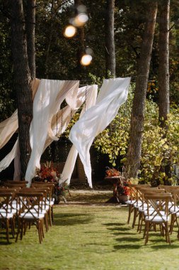 Dalgalı perdeler, ahşap sandalyeler ve ağaçlardan oluşan bir gölgeliğin altında canlı bir çiçek dekoru bulunan güzel bir açık hava düğün alanı..