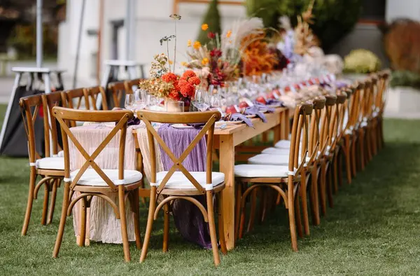 Canlı çiçeklerle süslenmiş uzun ahşap bir masa, zarif cam örtüler ve güzel katlanmış peçeteler içeren davetkar bir açık hava yemek düzenlemesi. Toplantılar için mükemmel..
