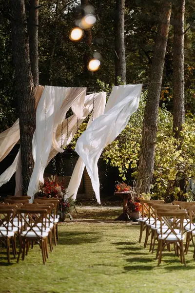 Dalgalı perdeler, ahşap sandalyeler ve ağaçlardan oluşan bir gölgeliğin altında canlı bir çiçek dekoru bulunan güzel bir açık hava düğün alanı..