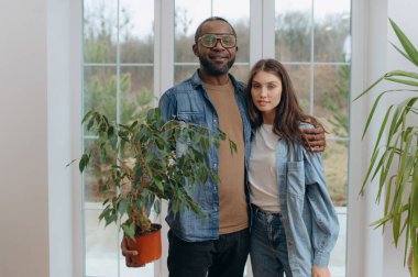 Neşeli bir çift parlak bir odada birlikte durur, bir saksı çiçeğini tutarken gülümserler, rahat bir ev ortamını gösterirler..
