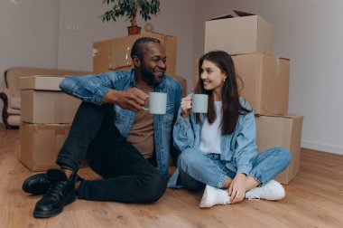 Neşeli bir çift, yeni evlerindeki kutuları taşırken yerde oturup kahveyi paylaşırken, yeni bir başlangıç sırasında sıcaklık ve bağlantı yayıyorlar..